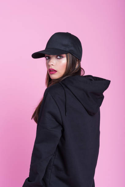 Young woman in black hooded shirt against pink background Studio portrait of beautiful young woman wearing black hooded shirt and baseball cap, standing against pink background and looking at camera. woman wearing baseball cap stock pictures, royalty-free photos & images