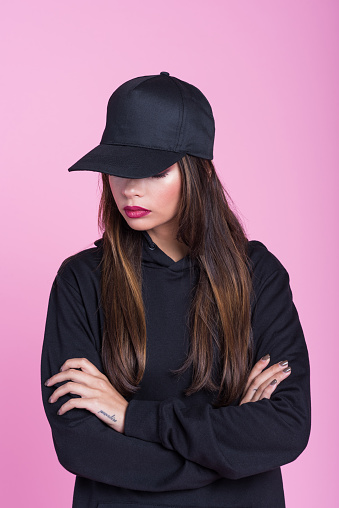 Studio portrait of sad young woman wearing black hooded shirt and baseball cap, standing with arms crossed against pink background.