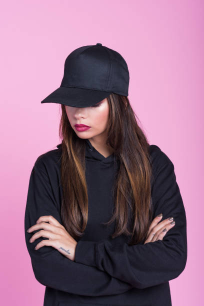Young woman in black hooded shirt and cap against pink background Studio portrait of sad young woman wearing black hooded shirt and baseball cap, standing with arms crossed against pink background. woman wearing baseball cap stock pictures, royalty-free photos & images