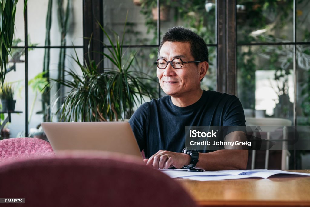 Smiling Chinese man working on laptop at home Mature man in glasses at home, typing on computer, contented, small business owner Men Stock Photo
