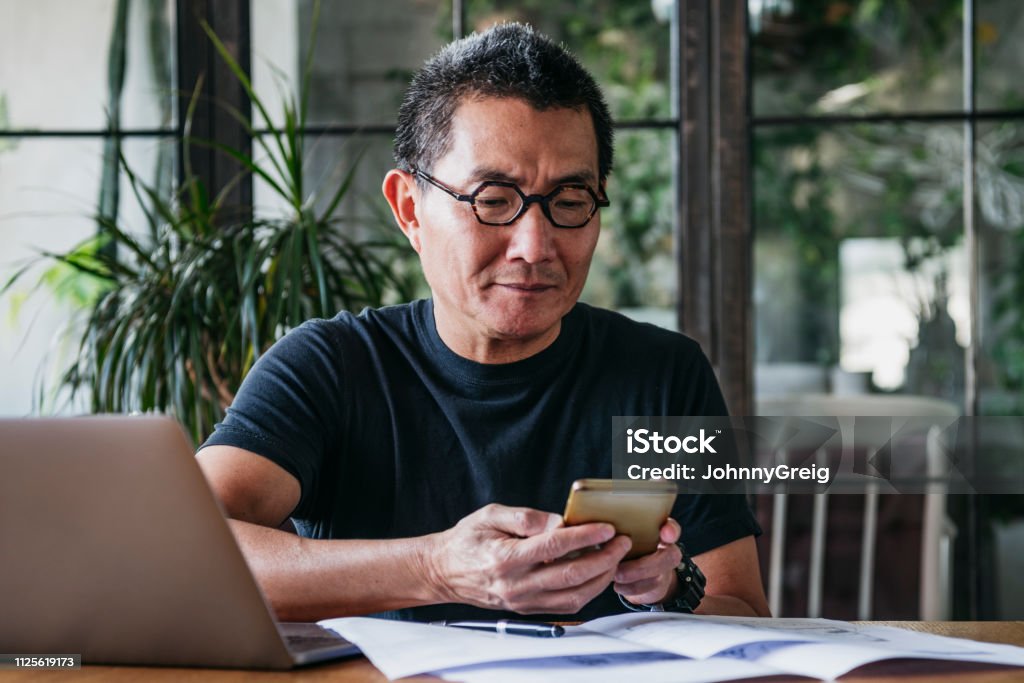 Mature man texting on cell phone with paperwork and laptop Chinese man in his 50s working remotely, using mobile phone, communication, connections, technology Financial Bill Stock Photo