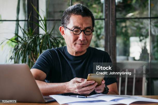 Mature Man Texting On Cell Phone With Paperwork And Laptop Stock Photo - Download Image Now