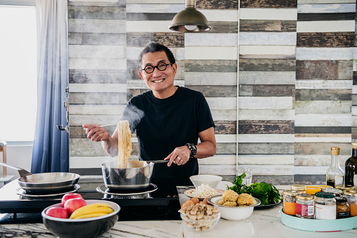 Man in his 50s holding saucepan with steaming noodles or spaghetti, smiling towards the camera