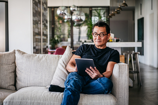 Chinese man in his 50s relaxing at home on sofa, casual clothes, reading ebook, surfing the internet