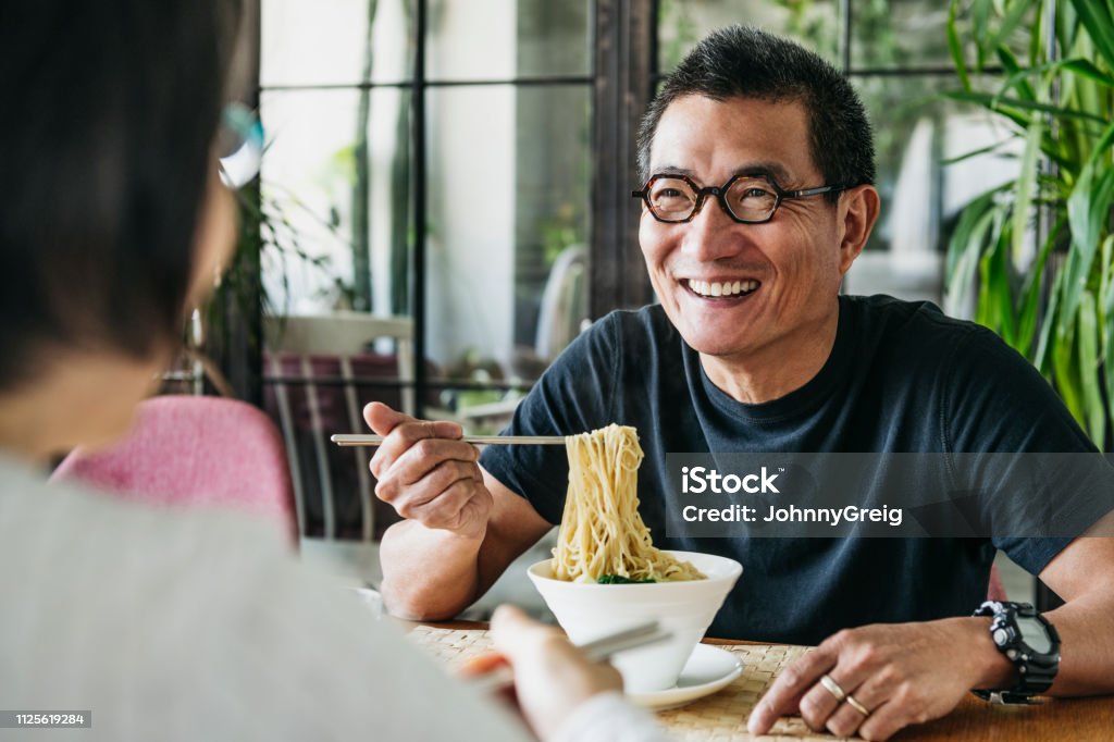 Mature man eating bowl of noodles and laughing Man in his 50s talking to woman and smiling, freshly made Chinese food, noodle soup, lunch, relaxation Eating Stock Photo