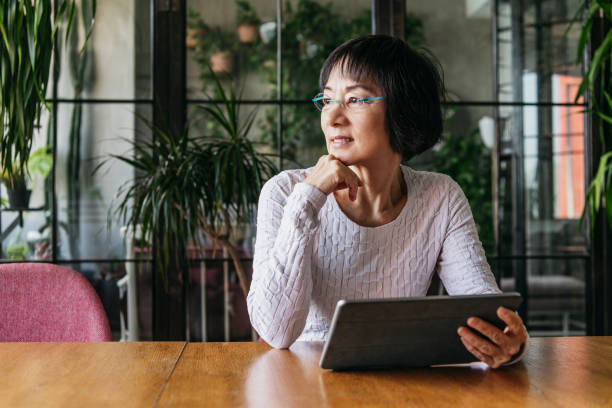 Chinese woman in her 60s using tablet with hand on chin Thoughtful senior woman sitting at home with digital device, looking away and daydreaming soul searching stock pictures, royalty-free photos & images