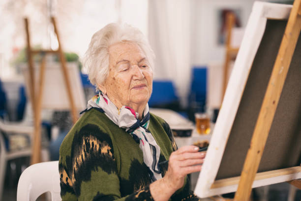 Close up senior woman with white hair painting on canvas in an art classroom. The woman sitting painting with a paintbrush.