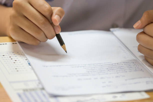 Education test concept : Man Hands high school, university student holding pencil for testing exams writing answer sheet and exercise for taking in exam paper on wood table at classroom with uniform