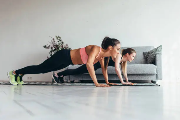 Two fitness women doing push-ups exercise working out at home. Side view.