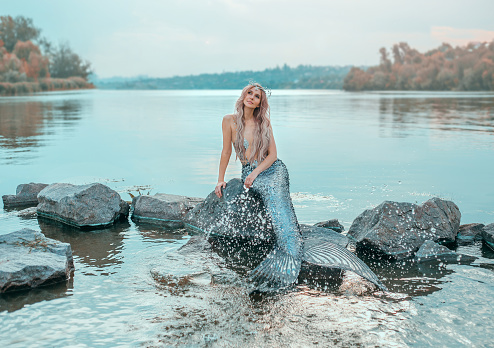 fair-haired mermaid in love dreams of handsome prince, new story Ariel, image of fairy-tale siren with long fish tail, chic crown of shells, plays, sprinkles clear water, ecology of environment.