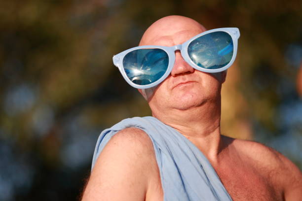 Humorous portrait of an adult bald man in a blue toga and funny blue glasses. On the beach, relax.