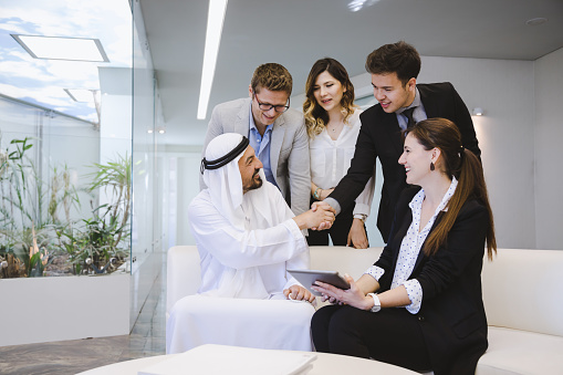 Young entrepreneur man with their colleagues presenting and shake hand about a new business projects and plans with digital tablet to poised and successful Emirati Arab Businessman in modern Office. UAE