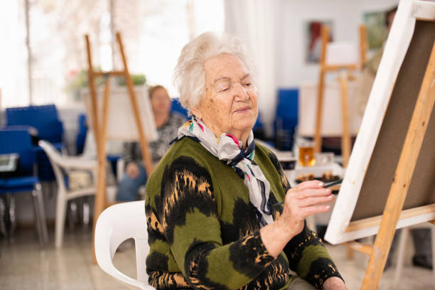 Senior woman with white hair painting on canvas in an art classroom. The woman wearing a green sweater, sitting on a chair, painting with a paintbrush. More classmates are in the background.