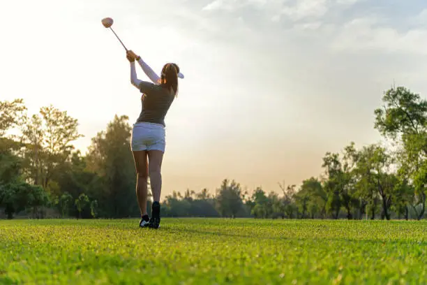 Photo of Sport Healthy. Asian sporty woman golf player doing golf swing tee off on the green sunset evening time, she presumably does exercise. Healthy and Lifestyle Concept.