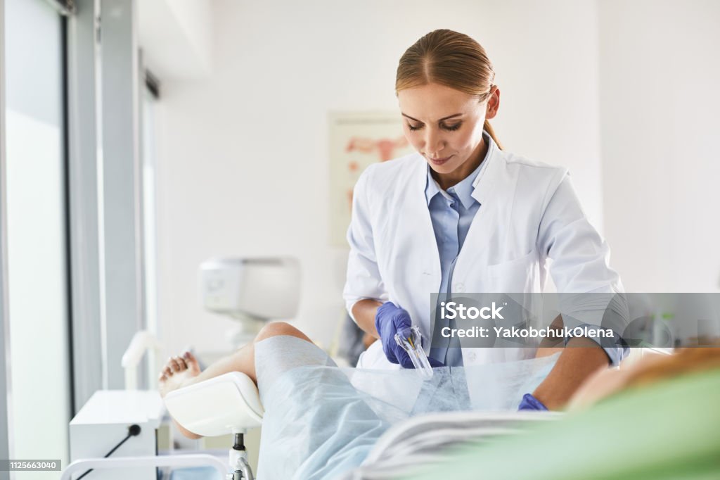 Young doctor doing gynecological examination of woman Waist up portrait of gynecologist in white lab coat and sterile gloves using vaginal speculum during pelvic exam. She is looking at instrument and smiling Gynecologist Stock Photo