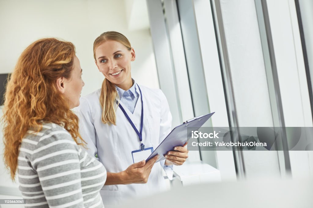 Young doctor holding clipboard while looking at patient Waist up portrait of smiling gynecologist standing with red-haired woman in medical office. Copy space in right side Doctor Stock Photo