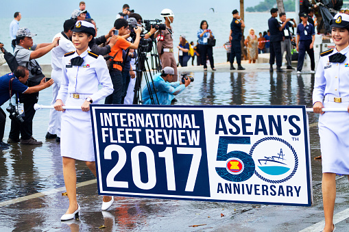 Female thai navy officers with Asean fleet parade banner are walking ahead of parade on street Beach Road in Pattaya. Parade for 50th anniversary of Asean's. In background are press and photographer media people.