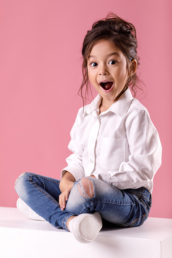 wow. Cute happy surprised little girl in white shirt with hairstyle looking to camera on pink background. Human emotions and facial expression