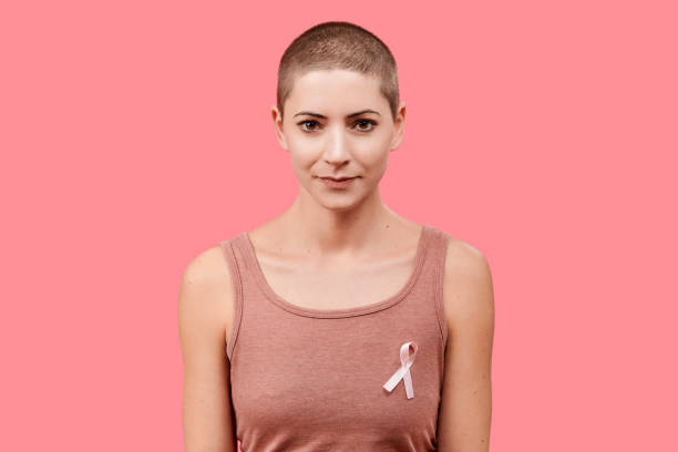 sourire mi 30 s femme, une survivante du cancer, portant un ruban de sensibilisation poitrine rose cancer, isolé au cours de la vie fond corail. concept de soutien, de solidarité, de dépistage et de prévention. - shaved head photos et images de collection