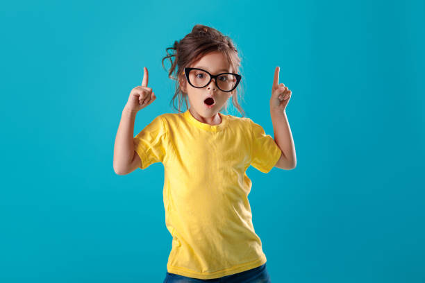 linda niña sorprendida en gafas y camiseta amarilla hacia arriba - child lifestyles isolated blue fotografías e imágenes de stock