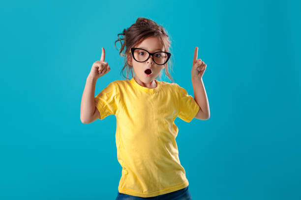 cute surprised little girl in glasses and yellow t-shirt pointing up - fashion model small one person happiness imagens e fotografias de stock