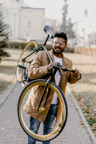 barbu de homme de hipster indienne avec un vélo à pignon fixe. young confiant barbu homme portant son vélo sur l’épaule et en regardant la caméra en se promenant à l’extérieur. homme d’affaires élégant avec un vélo. - caméra à lépaule photos et images de collection