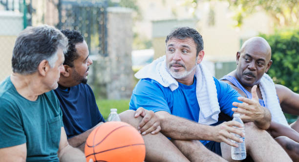 Group of men hanging out on basketball court, talking A group of four multi-ethnic mature and senior men in their 40s, 50s and 60s, taking a break after playing basketball. They are sitting on an outdoor court with water bottles and towels, relaxing, and talking. group of people men mature adult serious stock pictures, royalty-free photos & images