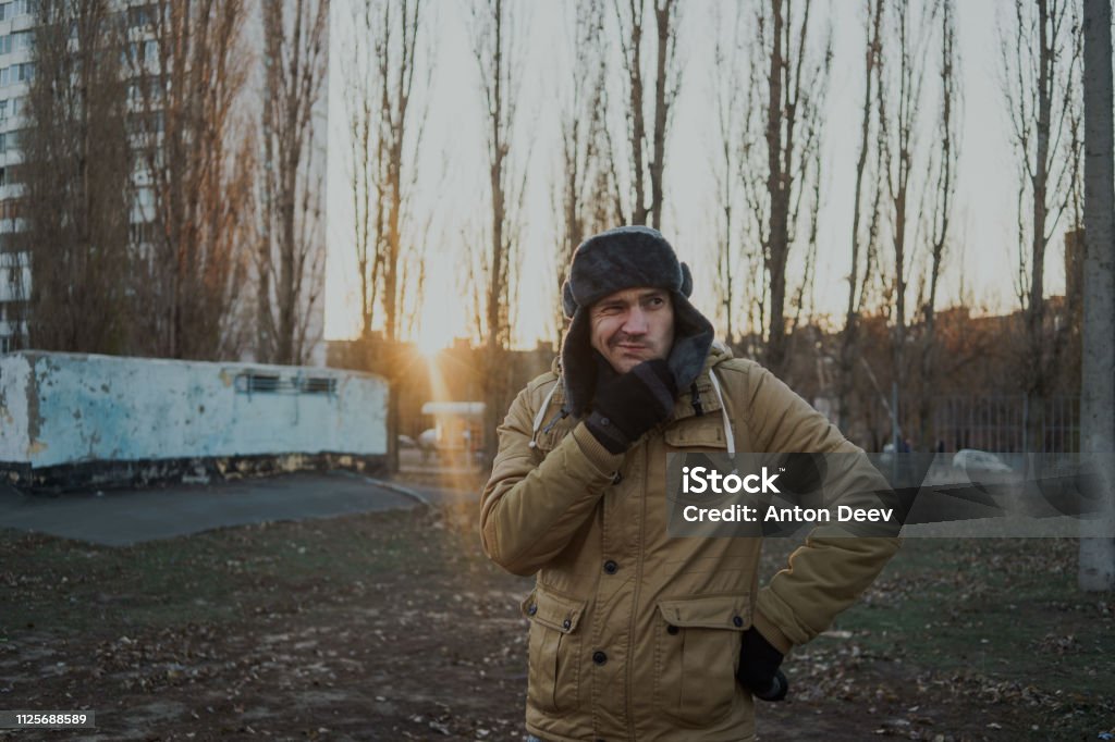 Happy handsome young man in cap with earflaps. The young man in the fur hat. a young guy standing on the street on a cloudy day. emotional portrait of a student. street style Adult Stock Photo