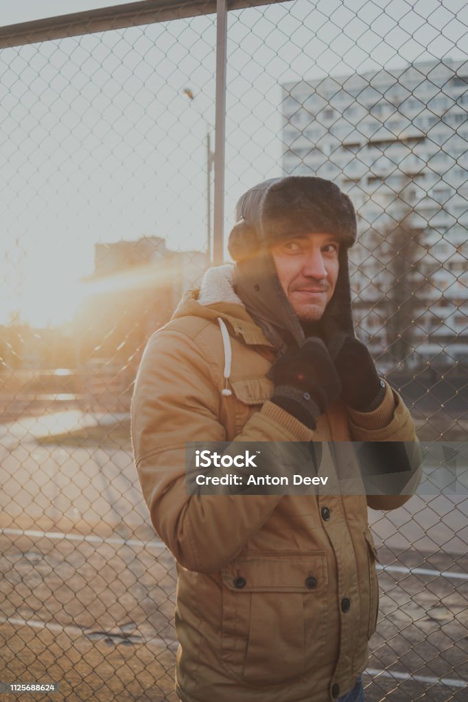 Happy handsome young man in cap with earflaps. The young man in the fur hat. a young guy standing on the street on a cloudy day. emotional portrait of a student. street style Adult Stock Photo