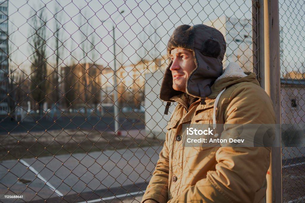 Happy handsome young man in cap with earflaps. The young man in the fur hat. a young guy standing on the street on a cloudy day. emotional portrait of a student. street style Adult Stock Photo