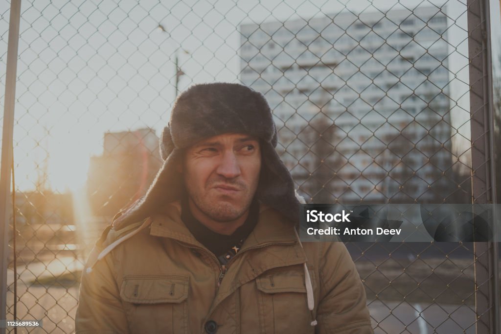 Happy handsome young man in cap with earflaps. The young man in the fur hat. a young guy standing on the street on a cloudy day. emotional portrait of a student. street style Adult Stock Photo