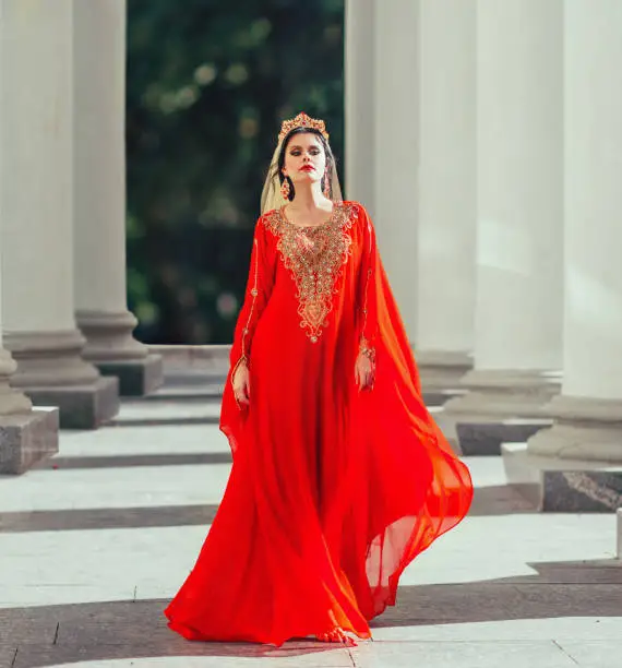 Gorgeous turkish empress shakherezad luxurious red flying dress with long train, sleeves, gold. walks smoothly between large columns with crown, earrings and bright scarlet lipstick. art sexy photo.