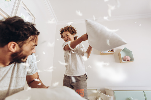 Happy father and son having a pillow fight on bed with feathers flying around. Father and son having fun playing at home.