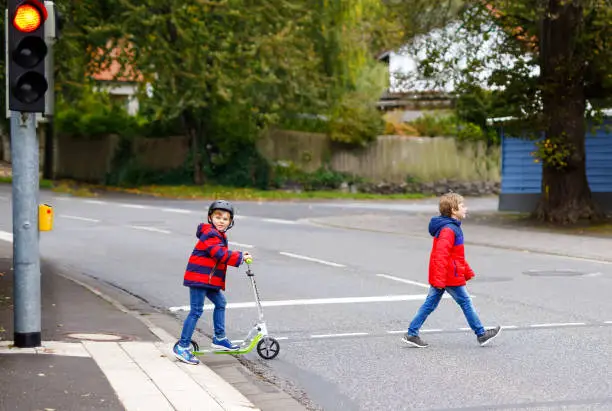 Photo of Two little schoolkids boys running and driving on scooter on autumn day. Happy children in colorful clothes and city traffic crossing pedestrian crosswalk with traffic lights.