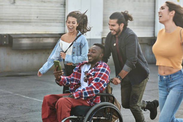 Man pushing friend on wheelchair while women running on street. Male and female hipsters enjoying city life. They are wearing casuals.