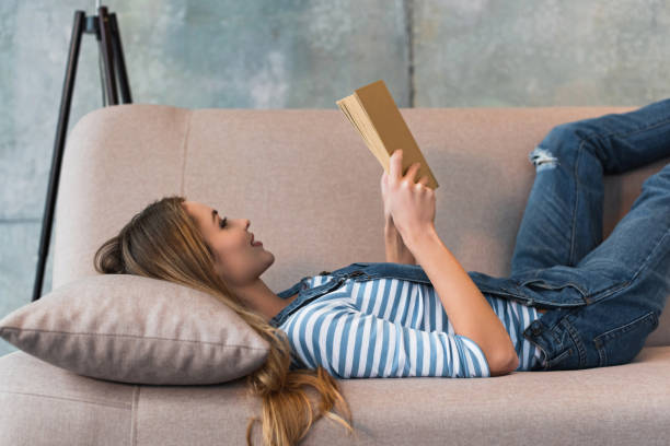 istock beautiful woman lying on sofa, smiling and reading book 1125698295