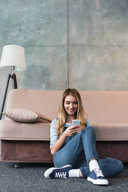 istock young girl smiling, using smartphone and sitting near sofa 1125698321