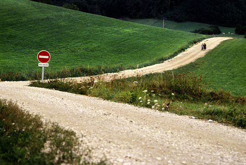 A curved road through the hills and a one way sign in the curve of the road. Two people are walking in the distance. Photo was taken in the Jura region in the eastern part of France.
