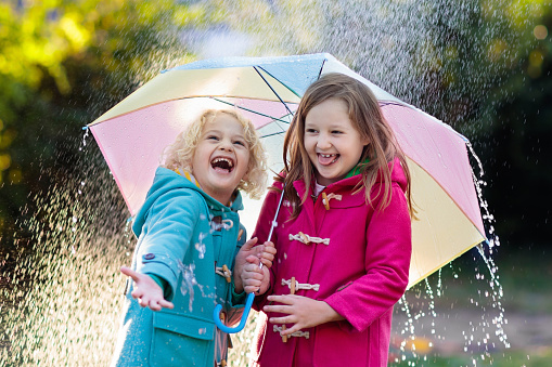 Kids with colorful umbrella playing in autumn shower rain. Little boy and girl in warm duffle coat play in a park by rainy weather. Fall outdoor fun for children. Kid catching rain drops.