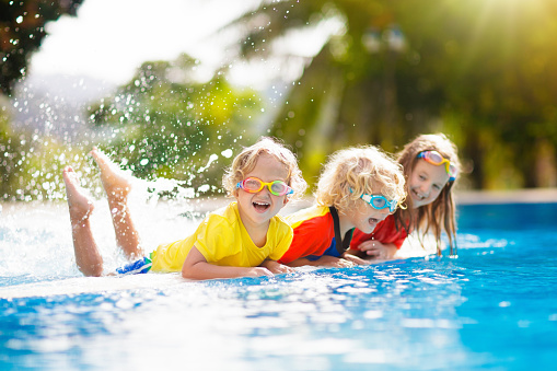 Kids play in swimming pool. Children learn to swim in outdoor pool of tropical resort during family summer vacation. Water and splash fun for young kid on holiday. Sun protection for child and baby.