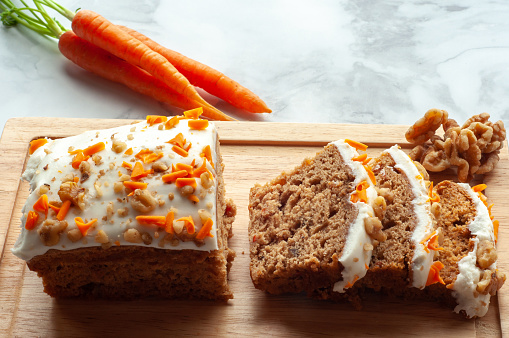 Carrot cake slices on wooden cutting board with whole carrots and walnuts in the background.
