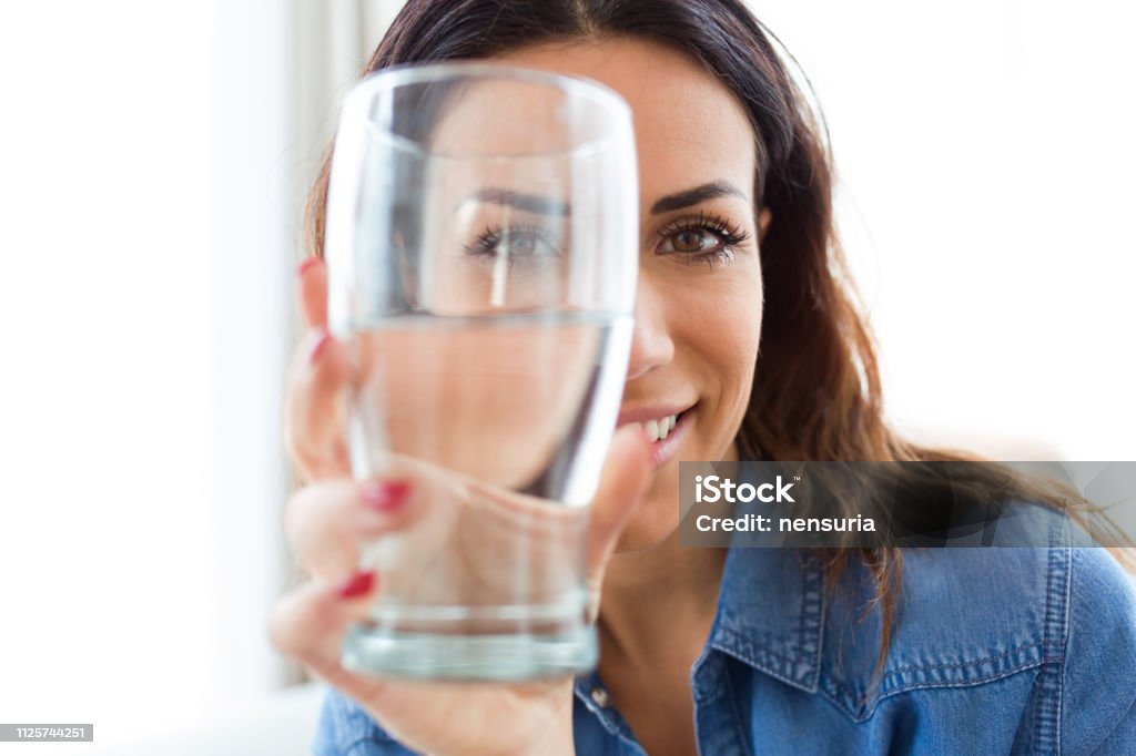 かなり若い女性が自宅の水のガラスを通してカメラを見ながら笑っています。 - 水のロイヤリティフリーストックフォト