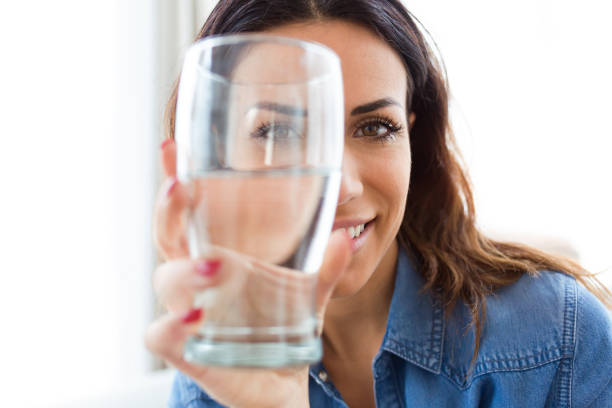 mooie jonge vrouw die lacht tijdens het kijken naar de camera via het glas water thuis. - drinking water stockfoto's en -beelden