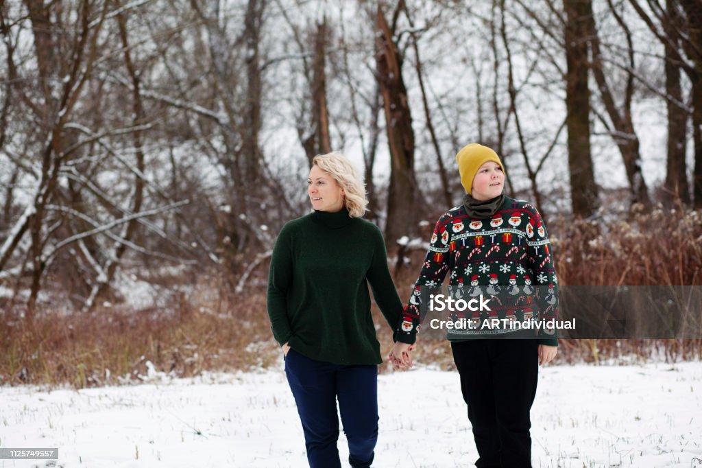 Beautiful mother and son hold hands while walking on snowy ground near to the forest. Warsaw, Poland - January 27, 2019: an attractive woman walks with her boy while looking away. Blond Hair Stock Photo