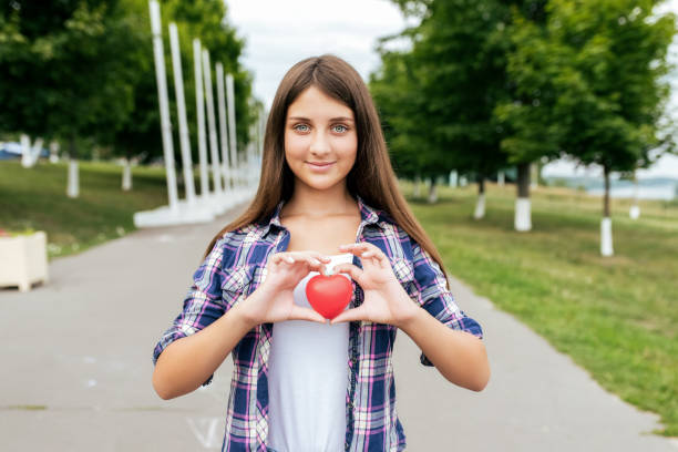 A teenager girl, 14-15 years old, stands in a city park in summer, holding a heart in her hands. In the summer, happy smiles. The concept of donation and transplant care for sick people. stock photo