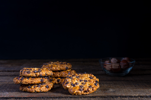 Some oats cookies with hazelnuts and chocolate chips on an old wooden table, a glass bowl with hazelnuts and dark background