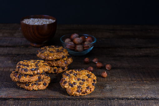 oats cookies with chocolate chips and hazelnuts on a wood table with a bowl of oats flakes and another one of hazelnuts with shell