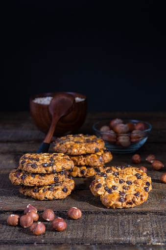 oats cookies with chocolate chips and hazelnuts on a wood table with a bowl of oats flakes and a spoon, another one of hazelnuts with shell