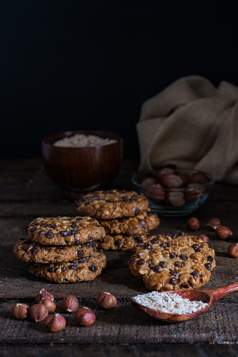 oats cookies with chocolate chips and hazelnuts on a wood table with a bowl of oats flakes and another one of hazelnuts with shell and a cloth