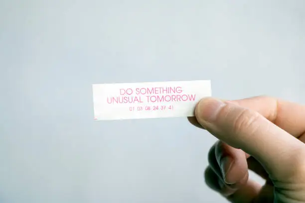 A hand holding a paper fortune from a fortune cookie reading, "Do something unusual tomorrow".
